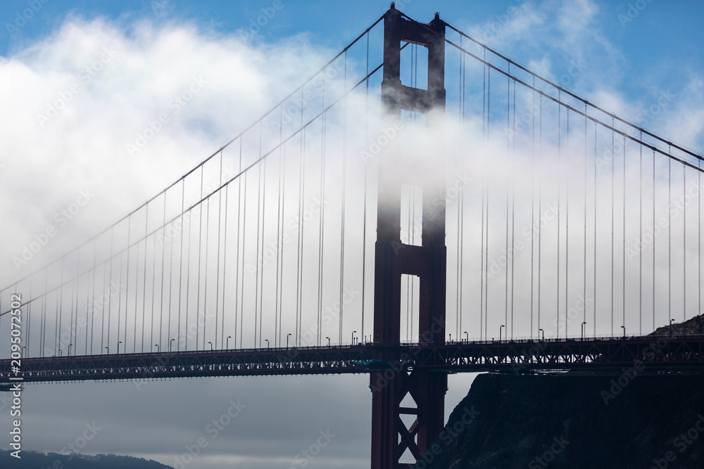 Fototapeta premium The Golden Gate Bridge is partially obscured by fog, showcasing its iconic towers and suspension cables against a backdrop of blue sky and clouds.