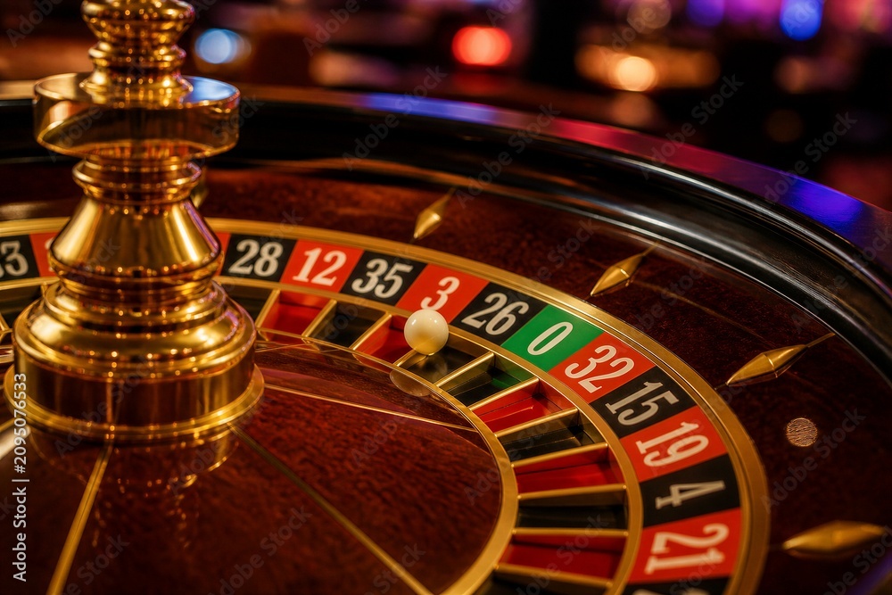Obraz premium The roulette wheel is in focus with a white ball nearing a number in the game. The background shows a busy casino filled with players and bright lights.