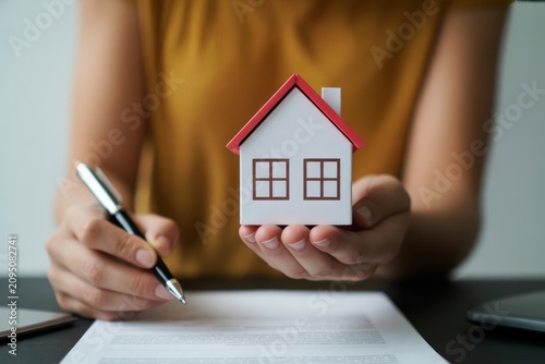 Person holding small white house model with red roof and pen on document