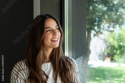 A smiling young woman with long brown hair gazes out a sunlit window in her home  appearing relaxed  hopeful and reflective. Natural light  casual clothing and a green backyard create a peaceful mood.