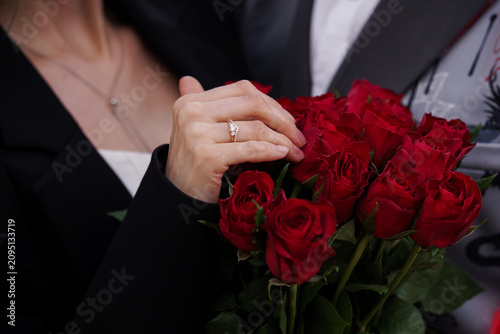 Close up of woman hand wearing engagement ring holding a bouquet of red roses in black formal attire