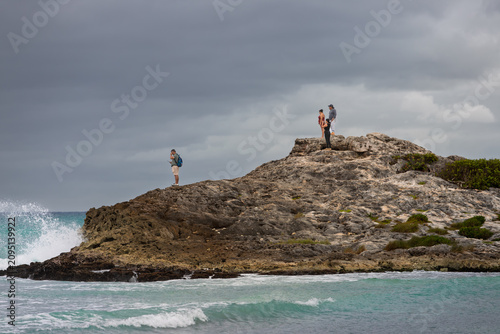 Tulum, Mexico - January 22, 2025: Tourists standing on a rocky limestone outcrop observing the crashing turquoise ocean waves under an overcast sky