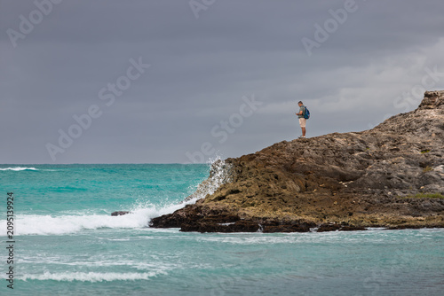 Tulum, Mexico - January 22, 2025: Tourist with backpack standing on a rocky limestone outcrop looking at his smartphone while ocean waves crash nearby