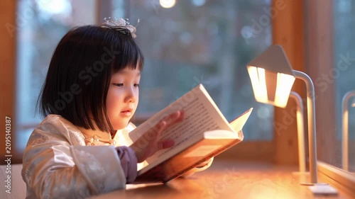 Soft light warms the room. Child reads quietly, eyes focused. Hands gently turn pages. Warm glow from lamp shines down. Cozy atmosphere, peaceful moment. Perfect for quiet study or bedtime scenes