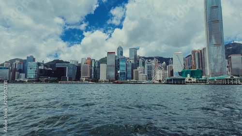 A sweeping panoramic view of Hong Kong's skyline from a water-based vantage point. The camera captures the dense urban landscape against the backdrop of towering skyscrapers