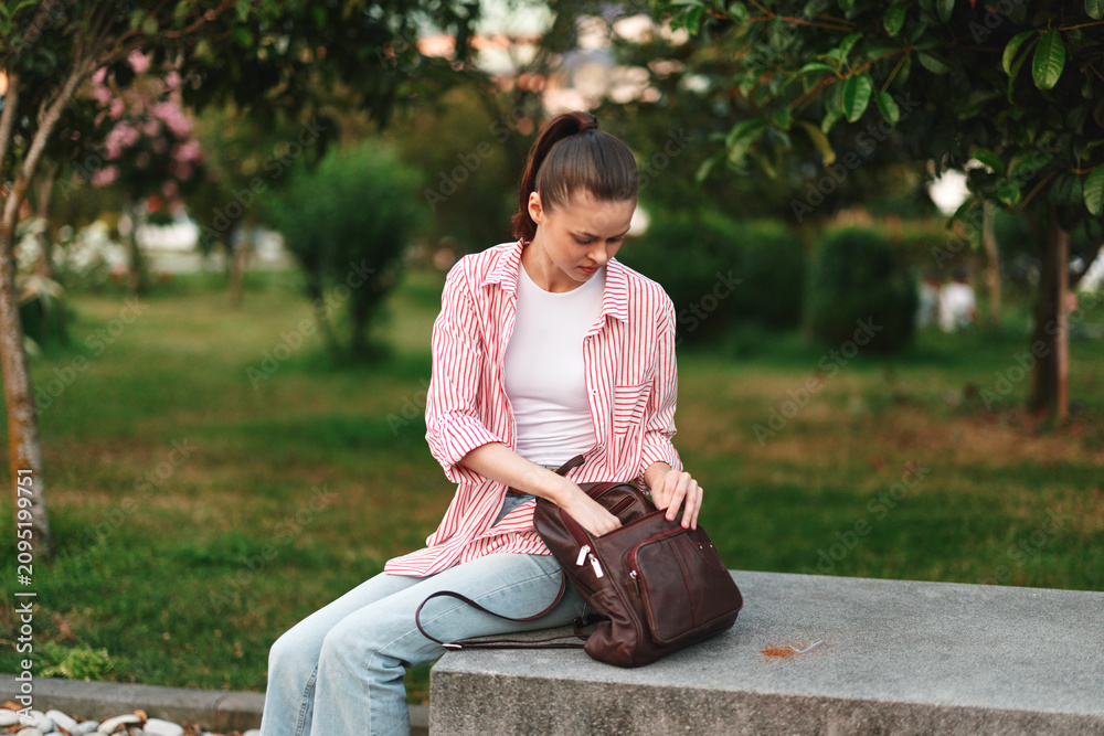Fototapeta premium Woman sits on a park bench with a backpack, wearing a striped shirt and jeans, enjoying a quiet outdoor moment in a green park setting.