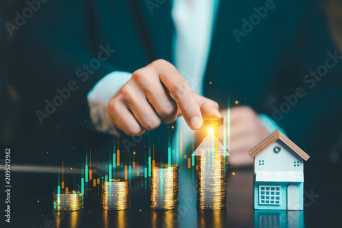 Close up of businessman hand stacking gold coins with growing stock market graph and small house model, real estate and financial concept.