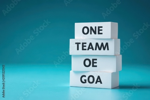 Four stacked white rectangular blocks on a teal surface with bold black letters reading ONE TEAM OE GOA, minimalist motivational concept and determined mood