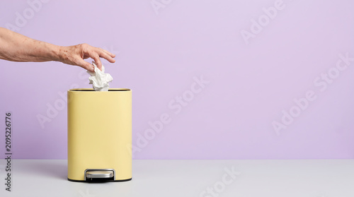 Elderly hand dropping crumpled white tissue into elegant trash bin on lavender background left side copy space