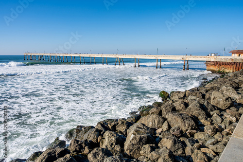 wide-angle view of churning white ocean surf crashing against a rocky shoreline next to a long public pier under a clear blue sky