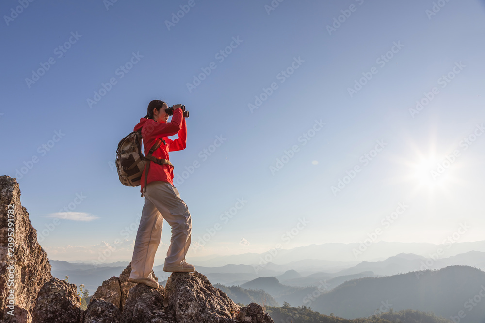Fototapeta premium Asian girl hiker with backpack holding binoculars standing on top of the rock mountain sunset sky background