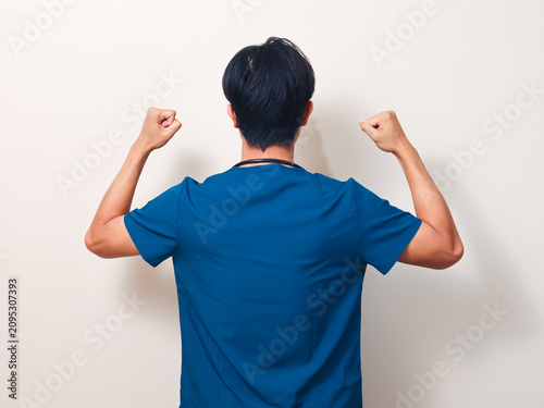 Rear view of a motivated young Asian male healthcare professional in a blue uniform celebrating with fists pumped against an isolated clean background.