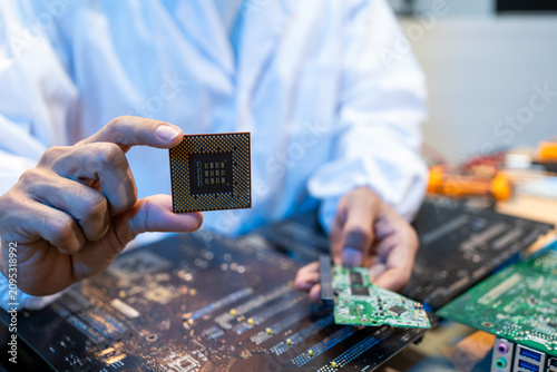 Technician in a lab coat holding a computer chip above circuit boards, showing electronics repair, hardware testing, and modern technology work in a close-up workspace scene.