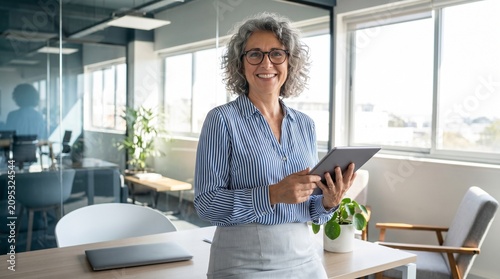 A modern office with glass walls and large windows; a person with gray curly hair and glasses stands in the foreground holding a tablet. On the desk are a closed laptop, notebook, pen.