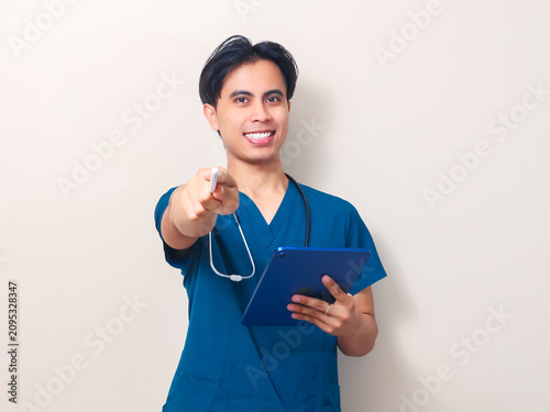 A smiling young Asian male healthcare professional in a blue uniform holding a digital tablet and pointing a stylus forward against an isolated clean background.
