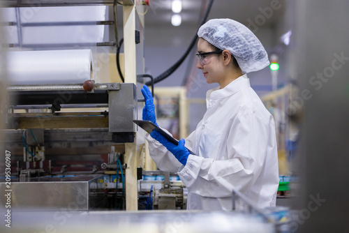 female worker inspecting manufacturing factory production line using digital tablet while wearing safety clean room uniform hairnet and blue protective glove showing attentive look