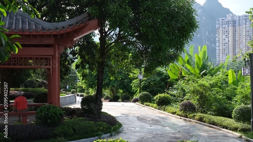 A senior man sitting or resting on a bench inside a classic Chinese wooden pavilion nestled within a lush public park. Cool, breezy refuge in a regional city. Local community, leisure space in Guangxi
