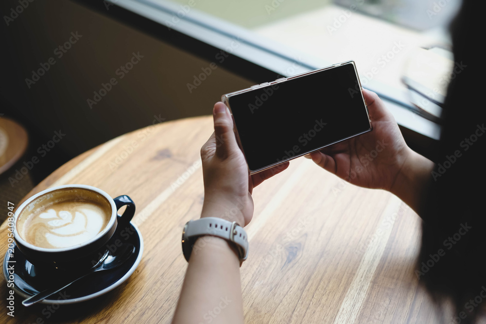 Obraz premium An over the shoulder view of a person holding a smartphone horizontally with a blank black mockup screen at a wooden cafe table next to a cup of coffee.