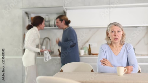 Upset old woman sitting in kitchen and man and woman arguing in background 