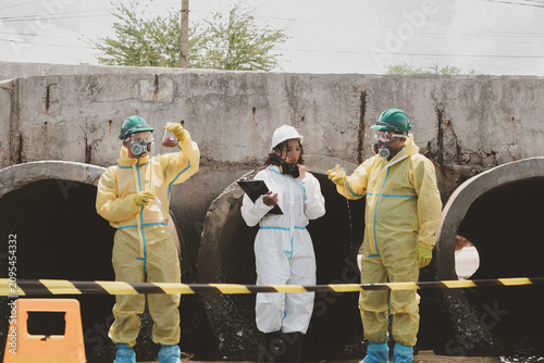 Scientists in chemical protective suits are at a water passage, collecting water samples and analyzing data on laptops to monitor pollution and contamination in the industrial area.
