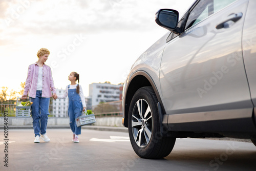 A Caucasian mother and her daughter walk hand-in-hand from a grocery store with shopping baskets towards their car