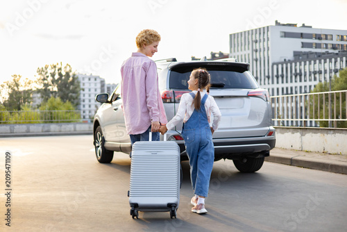 A Caucasian mother and her daughter hold hands while walking with a suitcase next to a car, ready for a journey