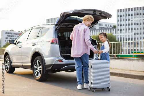 A Caucasian mother and her daughter are loading a suitcase into the back of a silver SUV parked on a city street