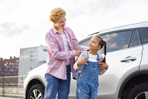 A smiling mother adjusts her daughter's backpack as they stand next to a car, preparing for a journey or school
