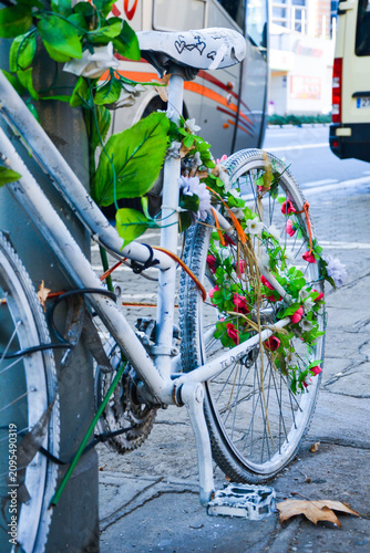 White bicycle adorned with green leaves and pink flowers parked against a pole on a city street, with blurred vehicles in the background