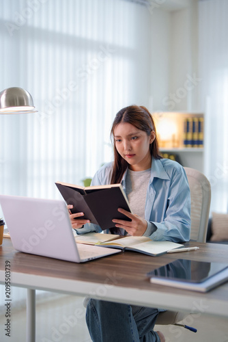 Young Asian student reading notebook while studying with laptop at home desk in bright interior