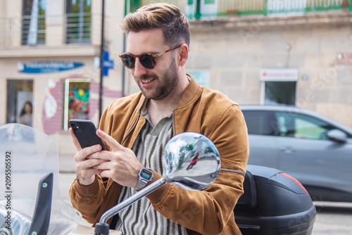 casual man sitting on motorcycle, smiling and texting on a mobile phone in the city