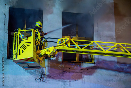 Quadro su tela Firefighter battling shopping mall fire from aerial ladder at night