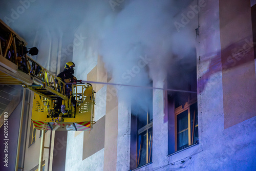 Quadro su tela Firefighter battling shopping mall fire from aerial ladder at night