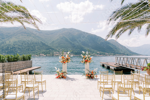 Elegant lakeside wedding ceremony setup with floral arch, chairs, palm trees, and mountain view