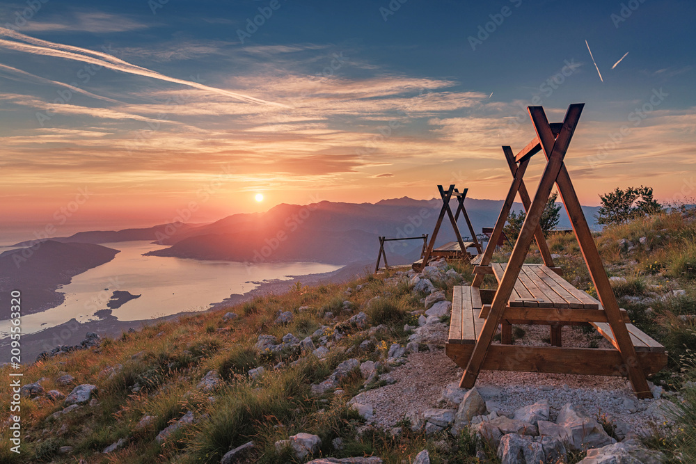 Naklejka premium Panoramic sunset illuminating the Bay of Kotor, Montenegro, from a mountain viewpoint with wooden picnic tables and benches