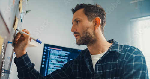Young Caucasian male programmer write creative plan on whiteboard and coding on computer in home office workspace. AI technology, cybersecurity network, agile strategy, business innovation concept.