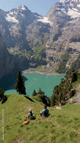 Oeschinensee Switzerland, is a breathtaking destination featuring turquoise waters surrounded by towering peaks. A couple of men and women relax on lush green hills, soaking in the beauty of nature.