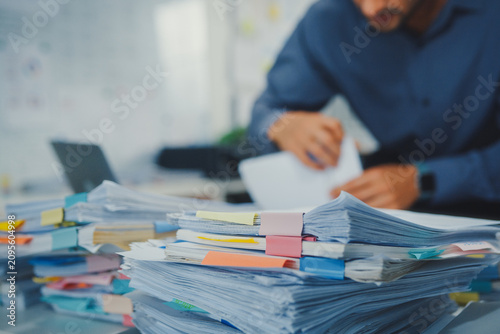 Foto Closeup overworked young Latin businessman reading document in large stack of paperwork file at office desk