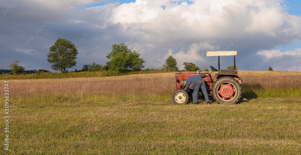 Obraz premium Farmer in workwear inspecting and repairing an old red tractor in a rural field, illustrating agricultural maintenance, mechanical problems and authentic countryside work