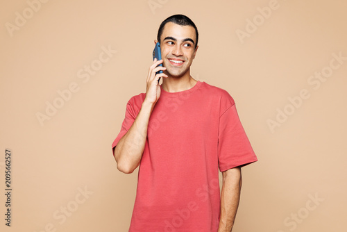 Young middle eastern man he wears red t-shirt casual clothes talk speak on mobile cell phone conducting pleasant conversation isolated on plain pastel light beige background studio. Lifestyle concept.
