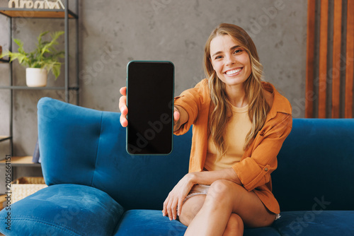 Young happy woman wear brown shirt hold in hand use mobile cell phone blank screen workspace sit on blue sofa stay at home hotel flat rest relax spend free spare time in living room indoors grey wall