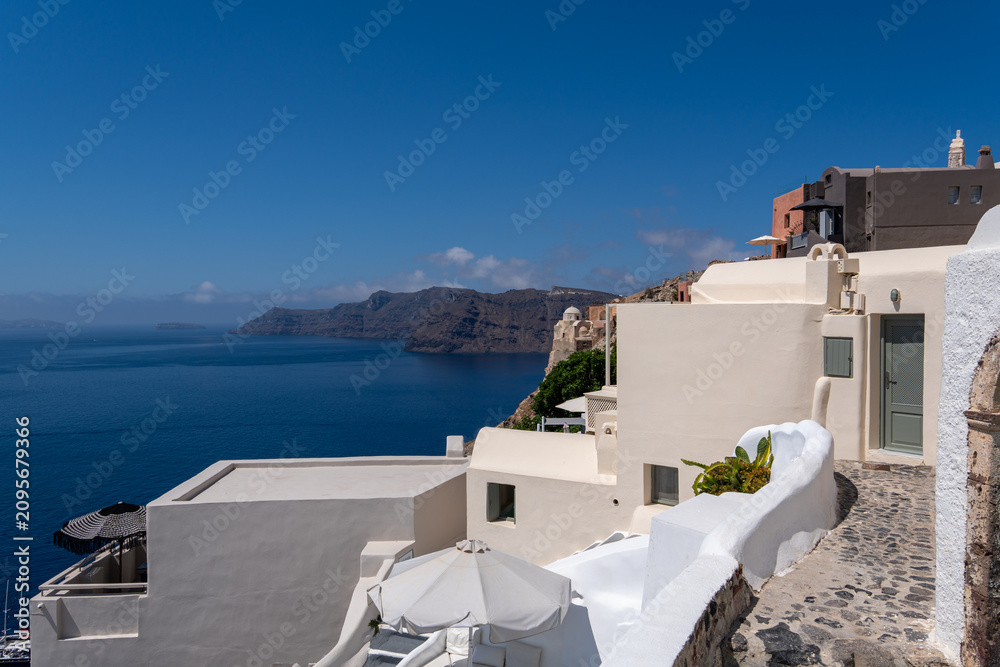 Fototapeta premium Traditional Cycladic architecture with white buildings and stone pathways on a cliffside in Oia, Santorini. Deep blue Aegean Sea and volcanic cliffs under a clear sunny sky.
