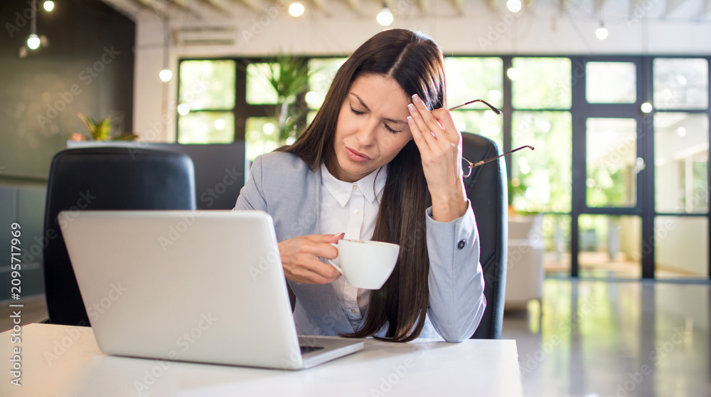 Fototapeta premium Stressed businesswoman holding cup of coffee and rubbing temple due to headache at work