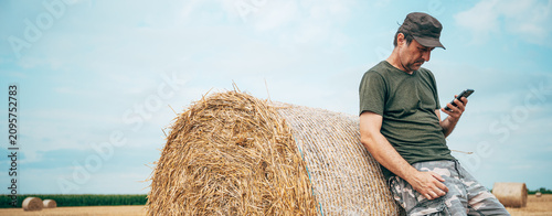 Farmer using a smartphone while leaning against a large round hay bale in a harvested field. Smart farming, digital agriculture and post-harvest farm management concept.