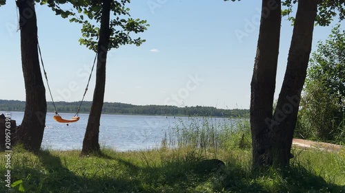  Empty yellow swing hanging between trees overlooking a calm blue lake with green grass and forest background on a sunny summer day.