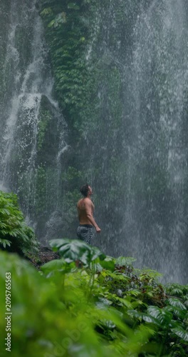 Handsome man with raised arms under powerful waterfall, feeling energy in tropical jungle nature
