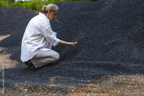 Fotografie Middle aged woman crouches beside large aggregate pile while collecting sample with one hand and holding a tablet