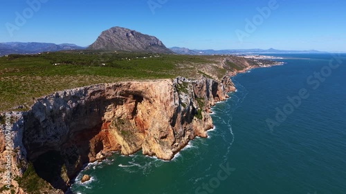 Aerial view of dramatic layered sandstone cliffs with a sea cave along the Mediterranean coastline near Javea, Alicante, Spain, with Montgo peak rising in the background