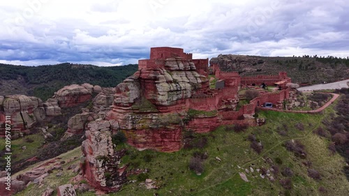 Aerial view of medieval Peracense Castle with crenellated defensive walls built atop dramatic red sandstone rock formations surrounded by green hills, Teruel, Aragon, Spain