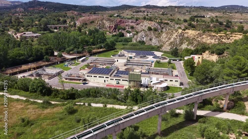 Aerial view of a solar-powered wastewater treatment facility nestled in a red rock canyon in Spain, with industrial conveyor bridge and historic stone viaduct visible among green trees